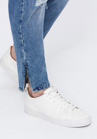 Zapatillas bajas blancas con parte superior de cuero liso, puntera redonda y suela de goma plana, combinadas con jeans azules desgastados que presentan cremalleras en los tobillos.