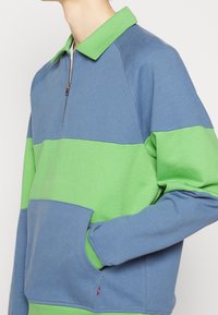 Pull-over bicolore avec col zippé. Comprend des panneaux bleus et verts. Confectionné en tissu texturé avec une poche avant et des poignets côtelés.