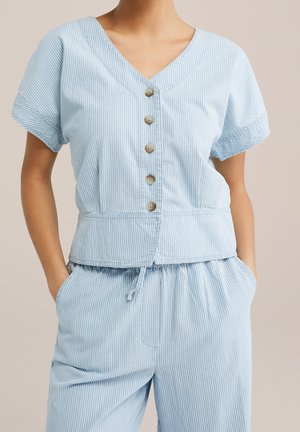 Femme portant un haut boutonné à manches courtes bleu clair et un pantalon à cordon assorti avec de fines rayures blanches verticales.