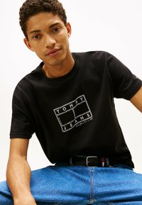 Zwarte katoenen t-shirt met een witte graphic "TOMMY JEANS" logo, gecombineerd met blauwe denim jeans en een zwarte riem met gekleurde accenten.