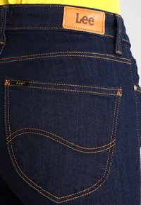 Marinblå denimjeans med en läder Lee-logotyp, kontrasterande orange sömmar och en rundad bakfickedesign.