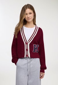 Giovane donna che indossa un cardigan lavorato a maglia bordeaux con strisce bianche e una grande lettera K, t-shirt bianca e pantaloni grigio chiaro con coulisse.