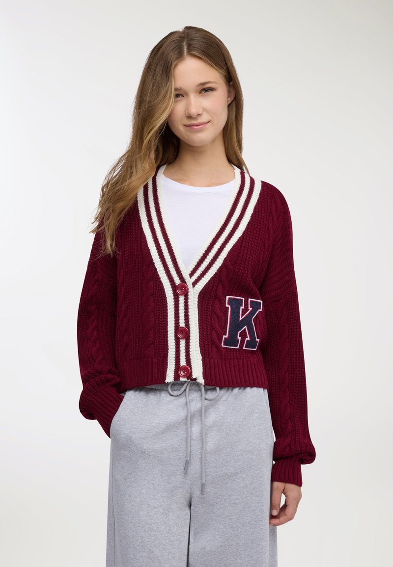 Giovane donna che indossa un cardigan lavorato a maglia bordeaux con strisce bianche e una grande lettera K, t-shirt bianca e pantaloni grigio chiaro con coulisse.
