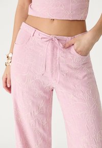 Pantalon rose brodé avec des motifs floraux, taille élastique à cordon, et un crop top assorti, main reposant dans la poche droite avec un bracelet au poignet gauche.