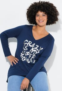 Marineblauwe T-shirt met lange mouwen en een V-hals, met metallic zilveren tekst "CRAZY IS MY SUPERPOWER." Zacht, casual model, gecombineerd met lichtblauwe jeans.