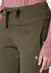 Olivově zelené tepláky s širokým, žebrovaným pasem, nastavitelným �šňůrkou a bočními kapsami. Na levé straně se nachází logo. Viditelný tenký, strukturovaný růžový top.