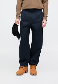 Luźne, ciemnoniebieskie spodnie wykonane z bawełny o gładkiej fakturze, zestawione z brązowymi zamszowymi sznurowanymi butami oraz czarną czapką trzymaną w ręku.