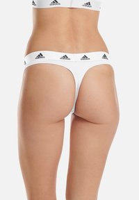 Weiße String-Unterwäsche mit einem breiten elastischen Bund, der das schwarze Adidas-Logo trägt. Glattes Material und minimale Rückabdeckung.
