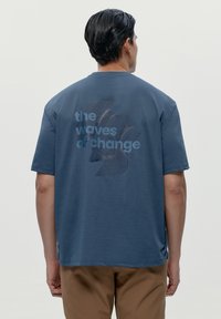 Modré bavlněné tričko s krátkým rukávem, na zadní straně s grafickým designem, který zahrnuje text "vlny změny".