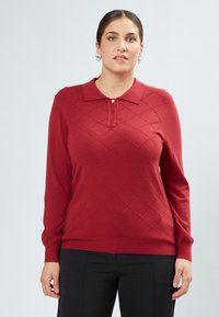 Pullover polo rosso a maniche lunghe realizzato in maglia, con un motivo a diamante, una patta con tre bottoni e polsini e orlo a costine. Abbinato a pantaloni neri.
