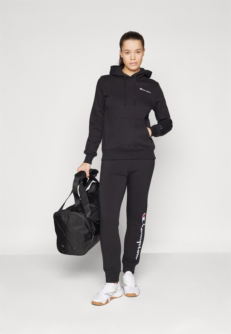 Schwarzer Hoodie mit Kängurutasche und Logo, kombiniert mit schwarzen Jogginghosen mit Logoprint. Begleitet von einer schwarzen Sporttasche.