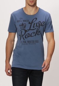 Modrá bavlněná tričko s kulatým výstřihem, na kterém je černý potisk textu: "Žij Rock" a "The Punk Monster Ball" s grafikou koruny.