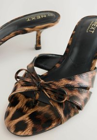 Mules korkeilla koroilla leopardikuvioisena, mustalla nahkaisella sisäosalla, etuosassa kierteinen rusetti-yksityiskohta ja hoikka stiletto-korko.