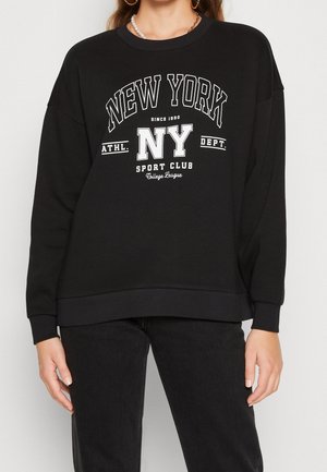 Femme portant un sweat-shirt noir avec le texte blanc "New York NY Sport Club College League", associé à un pantalon noir, des boucles d'oreilles anneaux dorés et un collier de perles.