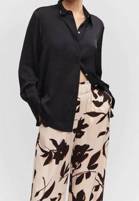 Chemise noire à boutons avec col, associée à un pantalon clair orné de motifs floraux sombres. Le tissu semble lisse et légèrement texturé.