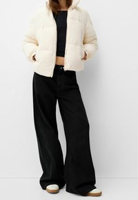 Giacca trapuntata crema con chiusura a zip e colletto a coste. Pantaloni neri a gamba larga. Scarpe da ginnastica bianche con suole marroni. Outfit semplice e casual.