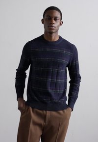 Sweater azul marinho com um padrão texturizado e riscas multicoloridas. Finalizado com um decote e punhos canelados. Combinado com calças castanhas.