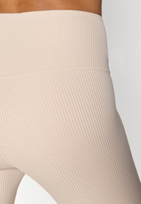 Beige rebraste tajice s širokim strukom, koje imaju mekanu teksturu i suptilan sjaj. Dizajn naglašava uski kroj.