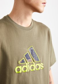 T-shirt en coton vert olive présentant un grand logo Adidas texturé jaune et violet avec trois accents rayés sur le devant.