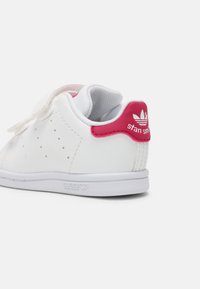 Vit lädersneaker med en rosa detalj vid hälen, med perforeringar på sidan, kardborreband och en texturerad gummisula.