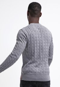 Pull gris à maille torsadée avec col rond, côtes texturées et ceinture ajustée. Comprend un détail en patch près de l'ourlet.