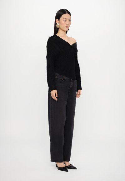 Black knitted off-shoulder sweater with textured pattern, paired with high-waisted dark jeans and pointed black heels. Gold earrings accent the look.