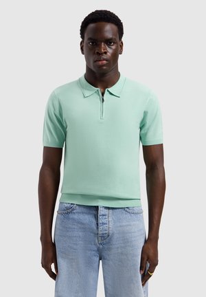 Mann steht, trägt ein mintgrünes kurzärmliges Poloshirt mit Reißverschluss am Kragen und hellblaue Jeans vor einfachem Hintergrund.