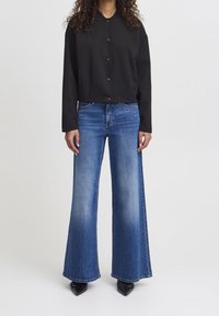 Czarna krótka kurtka z guzikami oraz szerokie niebieskie jeansy z wyblakłym wzorem i rozszerzanym krojem, w połączeniu z czarnymi, spiczastymi butami.