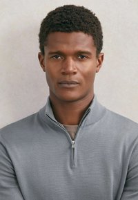 Młody mężczyzna z krótkimi kręconymi włosami, ubrany w szary sweter z suwakiem do połowy stójki, patrzący bezpośrednio w obiektyw na neutralnym tle.