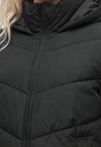 Gros plan d’une doudoune noire avec une fermeture éclair frontale et un col montant, portée par une personne aux cheveux blonds.