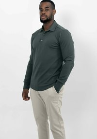 Donkergroene katoenen polo met lange mouwen, voorzien van een kraag, drie knopen en een subtiel logo. Gecombineerd met lichtbeige broeken.