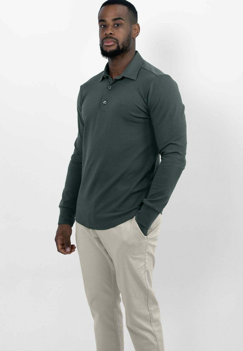Donkergroene katoenen polo met lange mouwen, voorzien van een kraag, drie knopen en een subtiel logo. Gecombineerd met lichtbeige broeken.