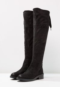 Paire de bottes hautes en daim noir avec talons bas carrés et bouts arrondis, debout sur une surface blanche devant un fond uni.