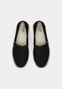 Svarta canvas slip-on skor med en texturerad espadrille-sula, med korsade design-accenter och en ljus inredning. TOMS-logotyp synlig inuti.