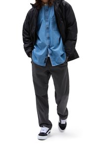 Hellblaues Jeanshemd mit Knöpfen, schwarze gesteppte Jacke, dunkelgraue Hose und schwarze Turnschuhe mit weißen Schnürsenkeln. Reflektierende Texturen vorhanden.