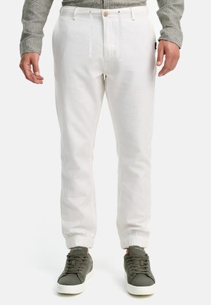 Mann trägt weiße Jogginghosen mit elastischen Bündchen, kombiniert mit grauen Sneakers und einem grauen Langarmshirt, steht gerade.