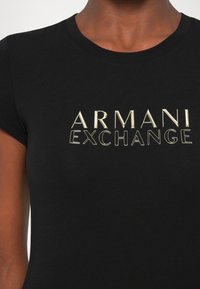Tricou negru din bumbac cu text „ARMANI EXCHANGE” auriu, realizat cu litere reliefate. Mâneci scurte și guler rotund. Textură netedă.