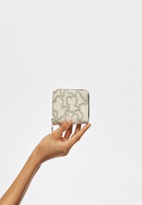 Cartera de crema texturizada con un diseño estampado en gris, que incluye un cierre de cremallera y un logotipo grabado en la parte frontal. Mano sosteniendo la cartera.