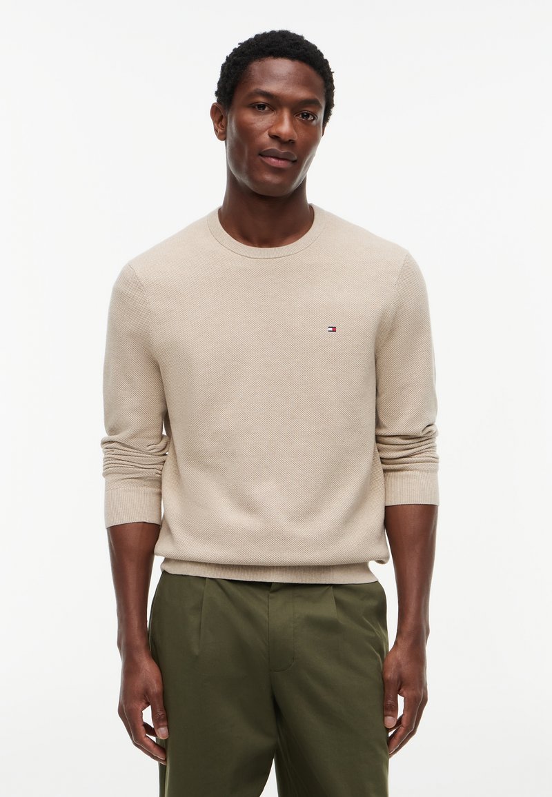 Homme portant un pull beige texturé à manches longues avec un petit logo sur la poitrine et un pantalon vert olive, debout sur fond blanc.