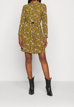 Kobieta w oliwkowej, zielonej sukience w kwiaty z długim rękawem i paskiem, w połączeniu z czarnymi botkami za kostkę z ostrym noskiem, stoi na tle jednolitego tła.