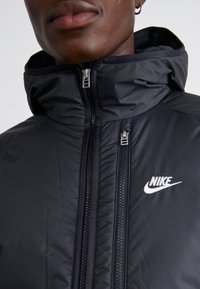 Czarna kurtka Nike z wysokim kołnierzem, z dwukierunkowym zamkiem błyskawicznym oraz logo Nike na piersi. Gładki, wodoodporny materiał.