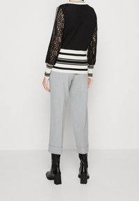 Vrouw die een zwart truitje met kanten mouwen en witte strepen draagt, lichtgrijze opgerolde broek, en zwarte enkellaarsjes met hakken, staand met haar rug naar de camera.