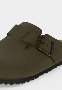 Καφέ δερμάτινο σανδάλι τύπου slip-on με μεγάλη μεταλλική αγκράφα και μαύρη ανάγλυφη σόλα.