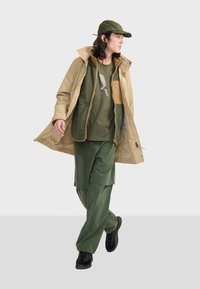 Giacca impermeabile beige con cappuccio, sovrapposta a un pile verde e pantaloni verdi. Sneakers nere, mentre cammina su uno sfondo neutro.