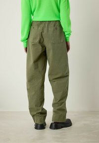 Alyvuogių žalios, laisvai krentančios kelnės su dviem užpakalinėmis kišenėmis ir tekstūruotu paviršiumi. Modelis vilki fluorescencinės žalios spalvos megztinį ir juodus batus.