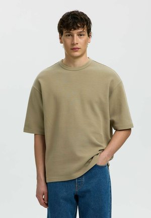 Junger Mann mit kurzen lockigen Haaren, der ein lockeres beiges kurzärmeliges, strukturiertes Hemd und blaue Jeans trägt, steht mit einer Hand in der Tasche.