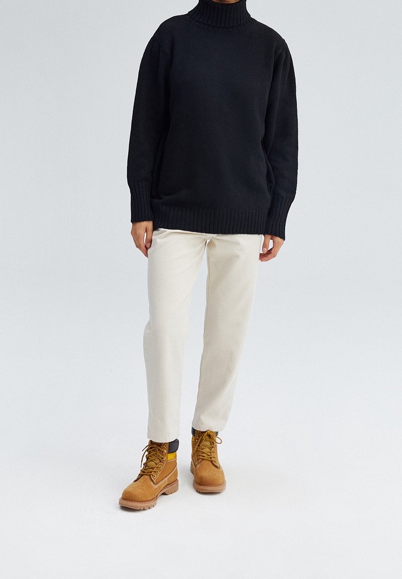 Czarny sweter na wysoki kołnierz, kremowe spodnie i brązowe buty z żółtymi akcentami. Sweter ma prążkowane krawędzie; spodnie są gładkie.