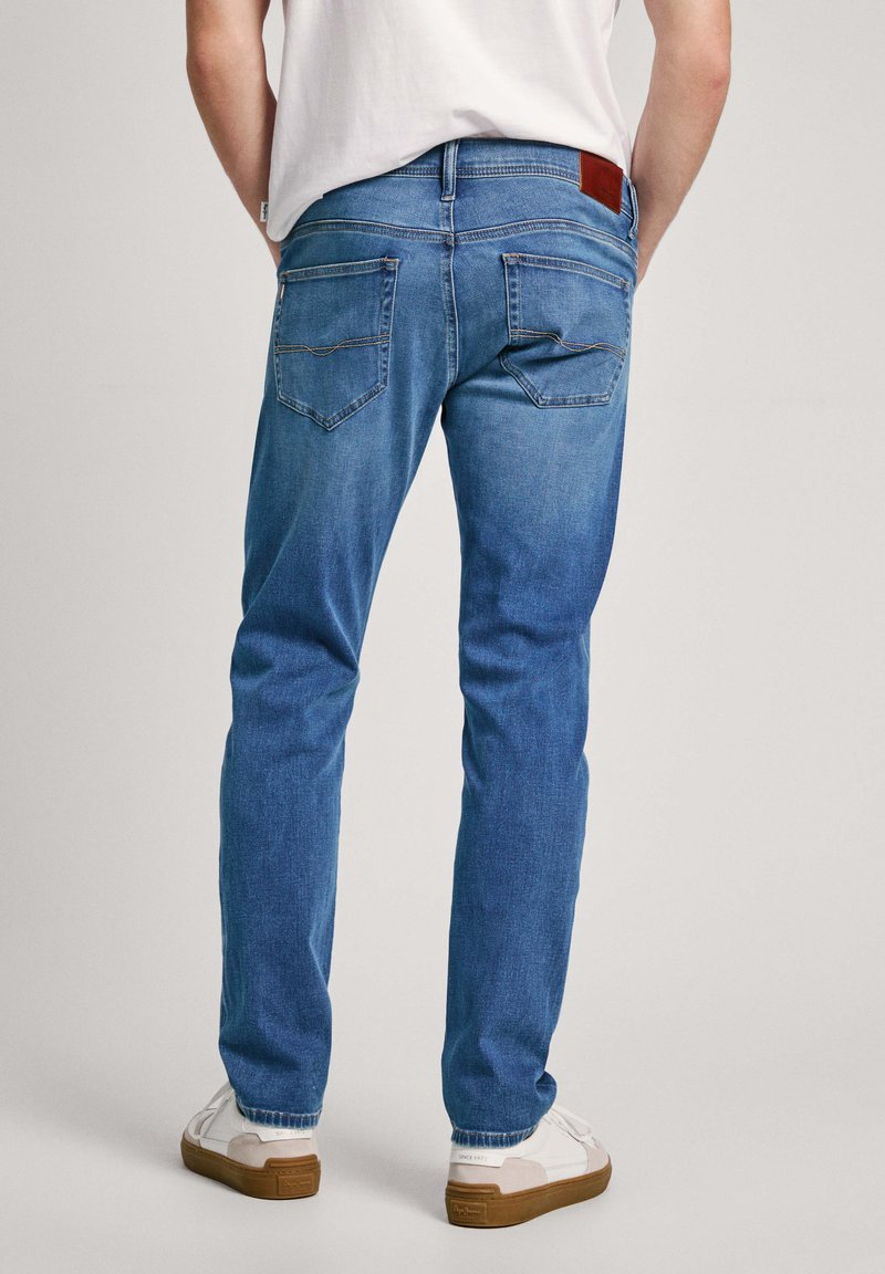 Vaqueros de denim azul con corte recto, diseño de cinco bolsillos, parche trasero de cuero y detalles de costura visibles. Combinados con zapatillas blancas.