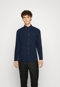 Chemise bleu marine à boutons, à manches longues, avec col, poche avant et boutons argentés ; texture lisse et coupe ajustée.