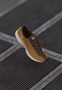 Rjave in bež sneakersi z perforacijami, rjavimi vezalkami in belim podplatom; na jezičku je Nike blagovna znamka. Imajo teksturirano površino in poenostavljen dizajn.